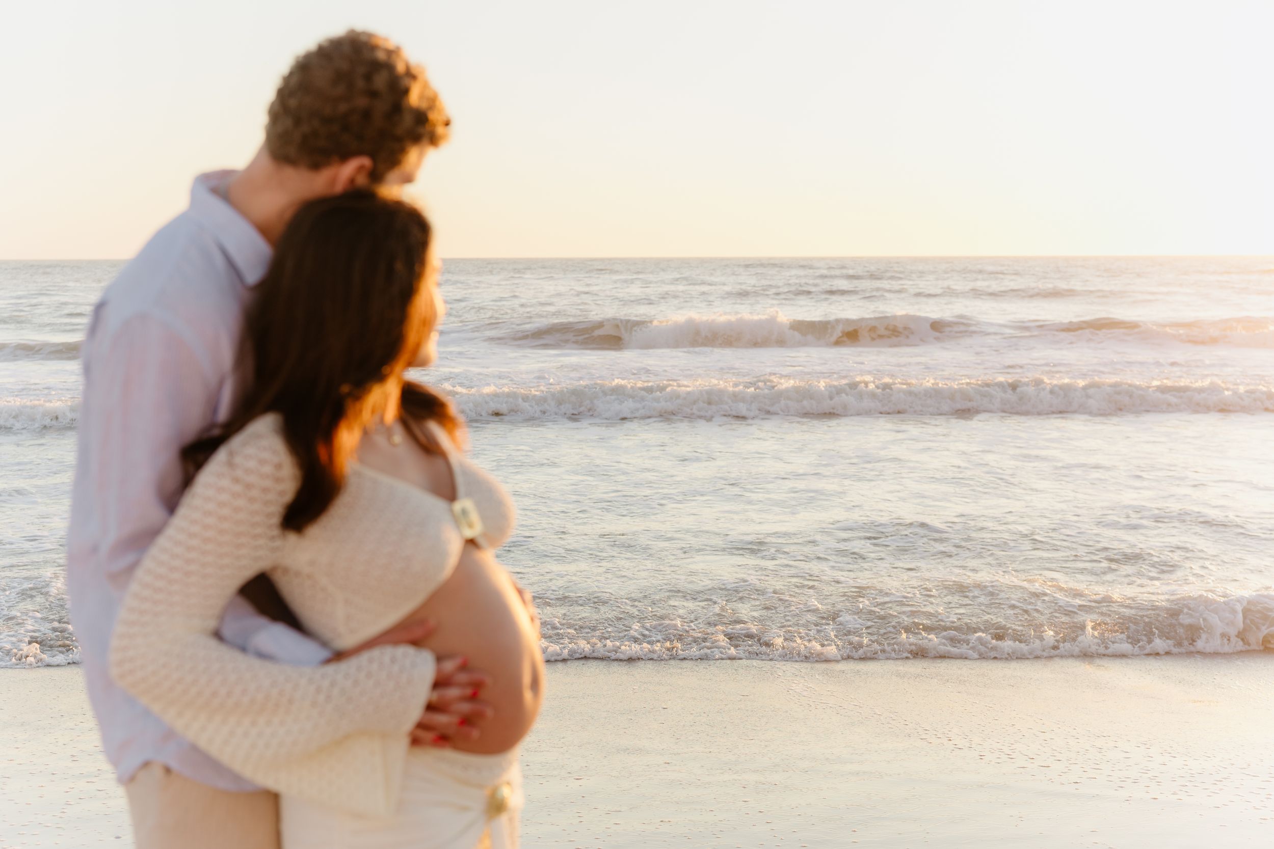 A couple embraces on a sandy beach at sunset with ocean waves in the background.