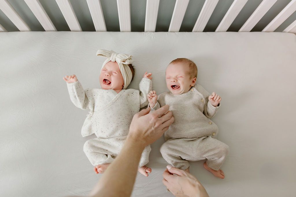 Two crying newborn twins in white onesies lying in a crib while a parent reaches in to comfort them.