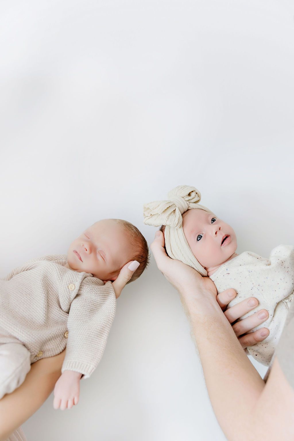 Two newborn twins lying side by side, one sleeping and one awake wearing a bow headband, on white background.