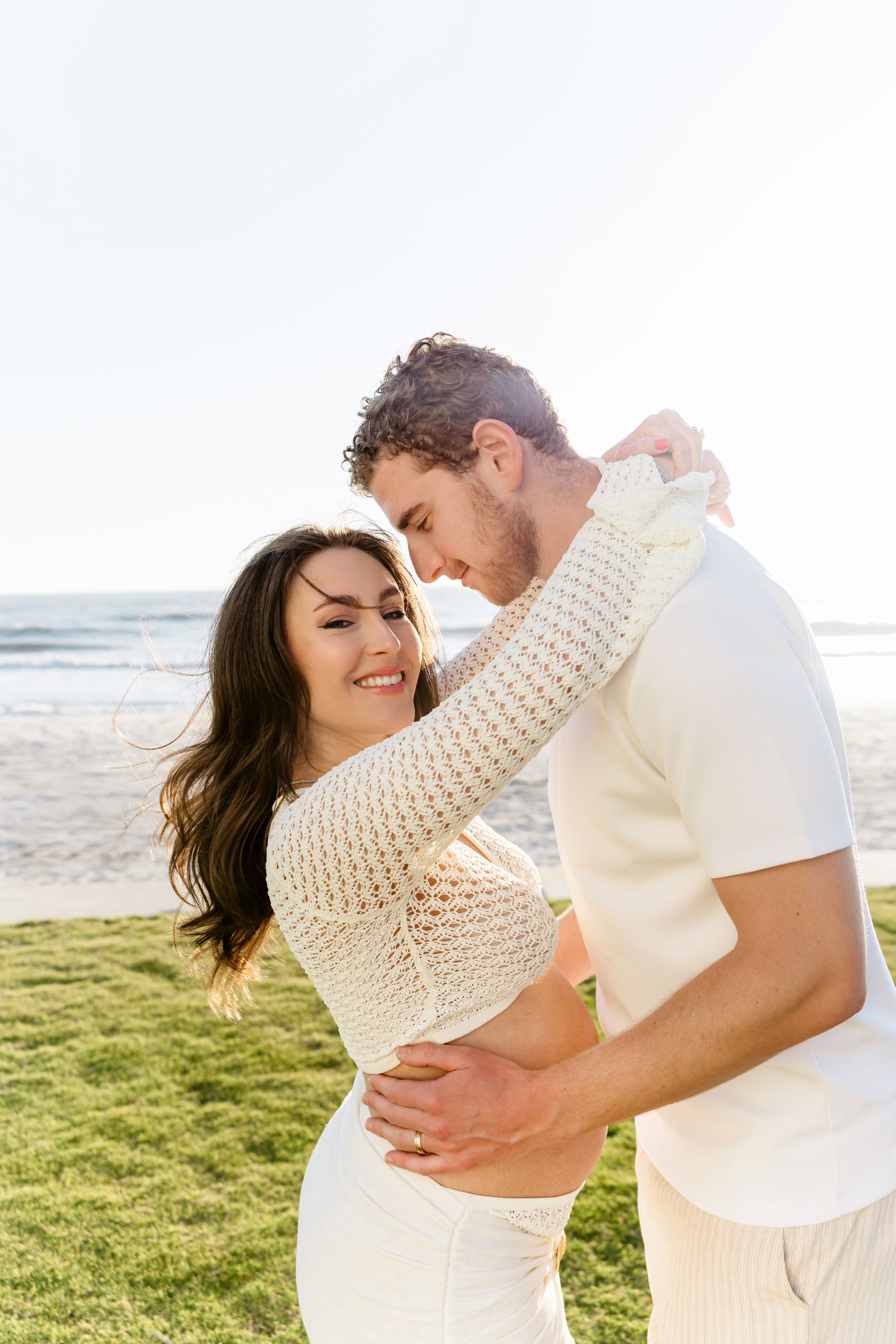 A romantic couple embraces and shares intimate moments on a sunny beach with grass and ocean views.