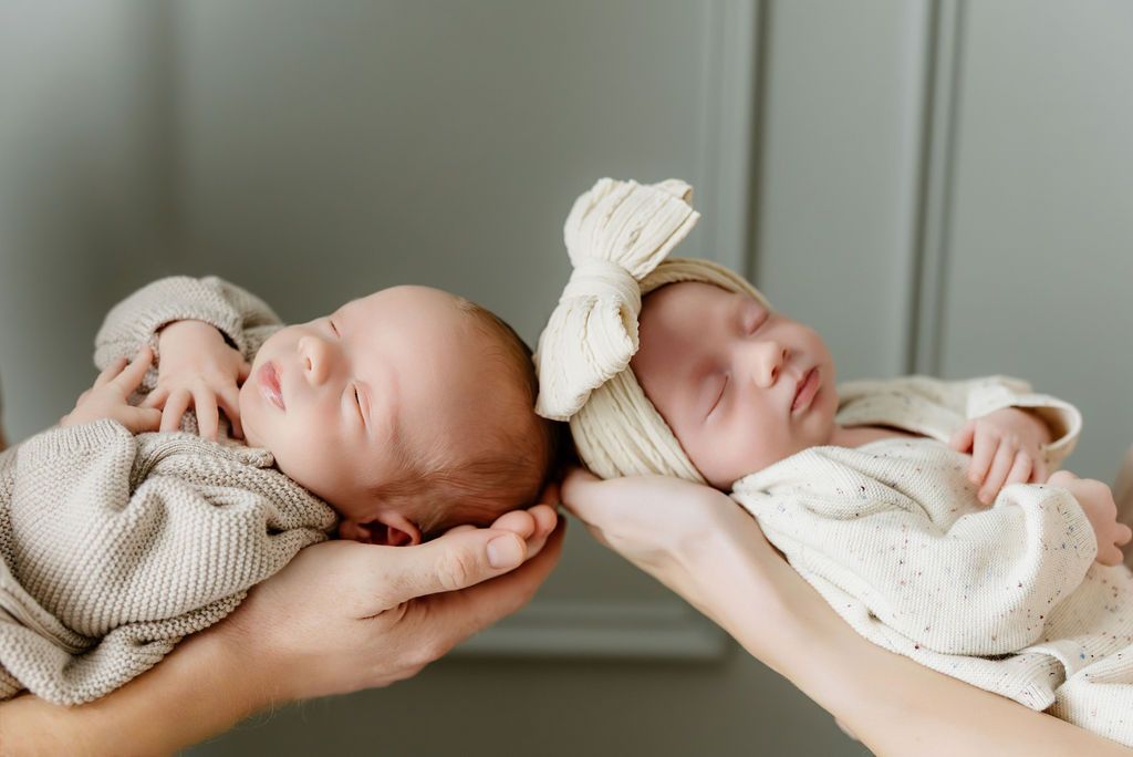 Two sleeping newborn twins held in hands, wearing soft beige outfits, one with a bow headband, in a serene studio setting.