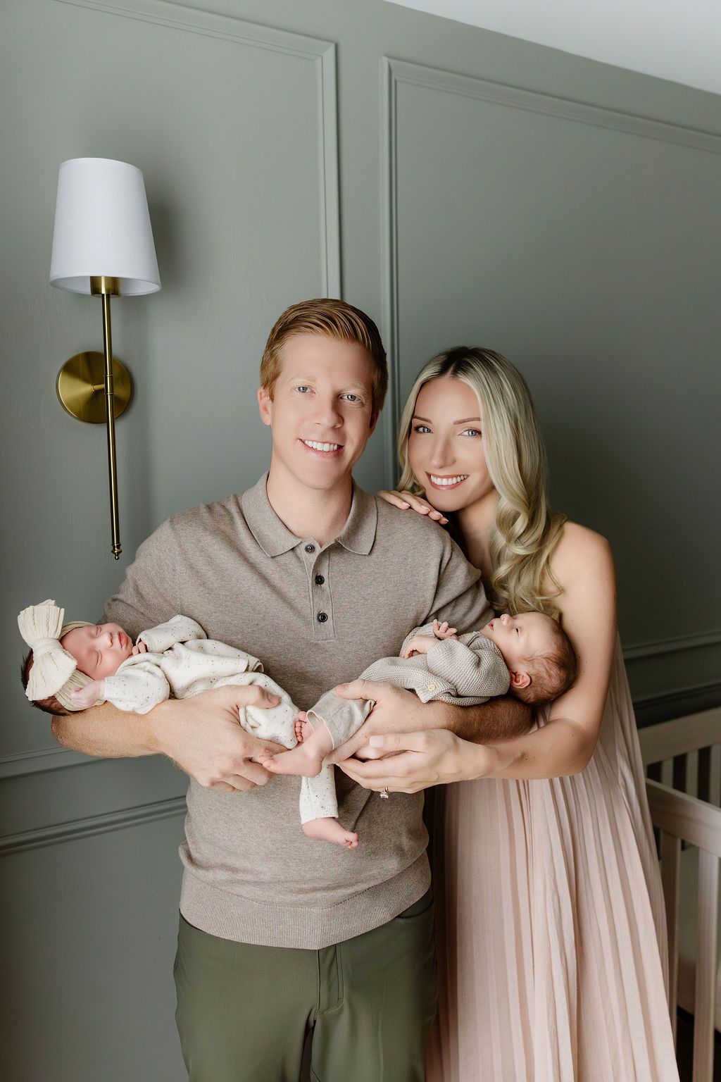 Smiling couple holding twin newborn babies in a nursery with sage green walls and a gold wall sconce.