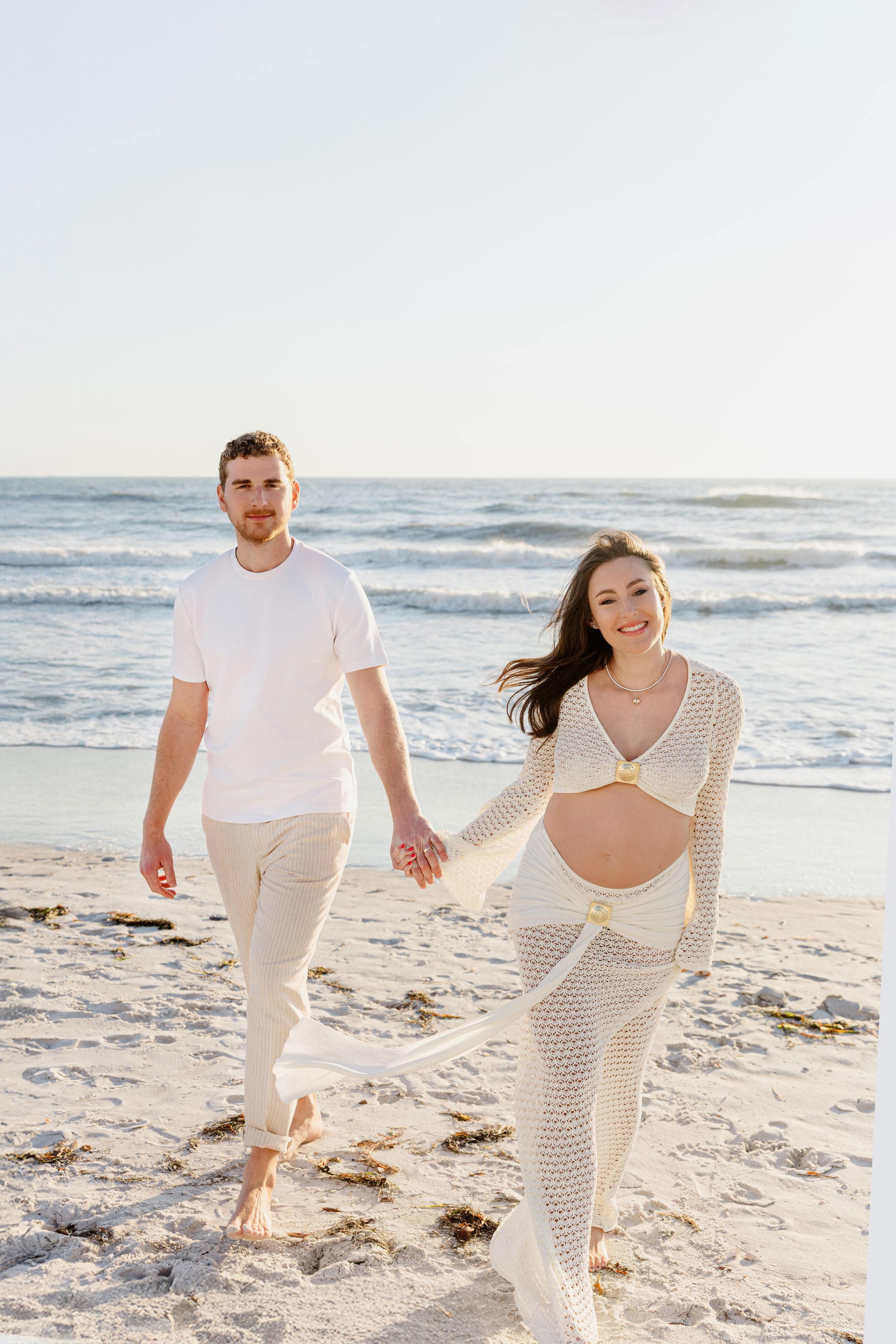 A couple in white outfits walks together on a sunny beach with ocean waves in the background.
