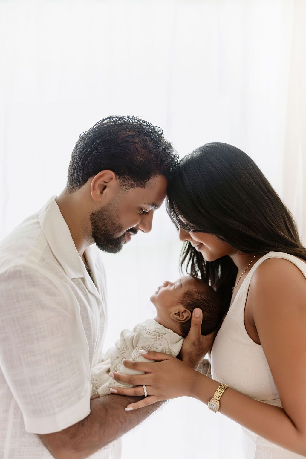 Two parents tenderly hold their sleeping newborn baby together, touching foreheads in an intimate family portrait.