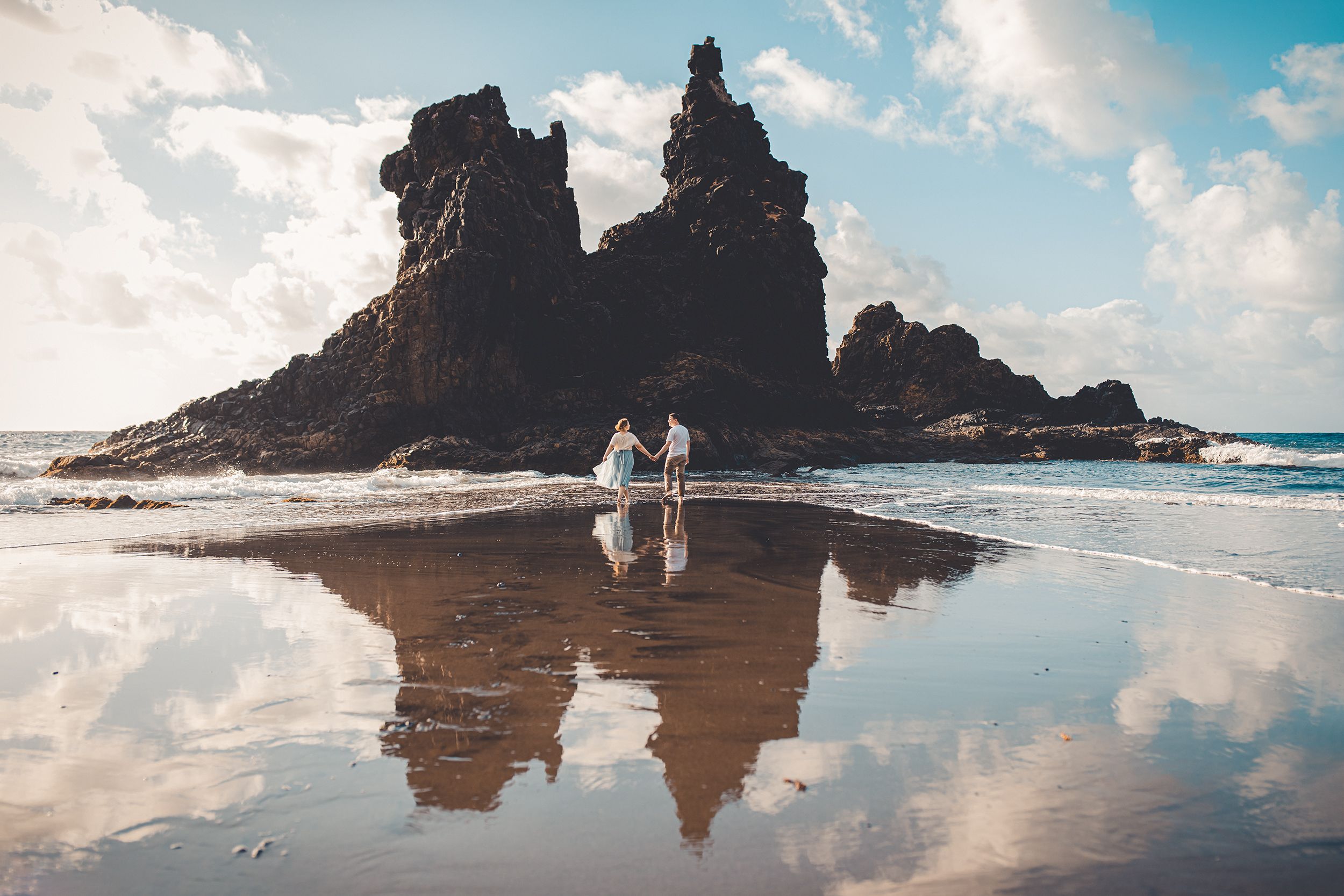 A couple holds hands, walking on a beach with rock formations and reflections, under a sunny, cloudy sky.
