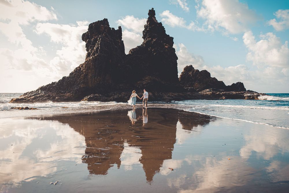 A couple holds hands, walking on a beach with rock formations and reflections, under a sunny, cloudy sky.