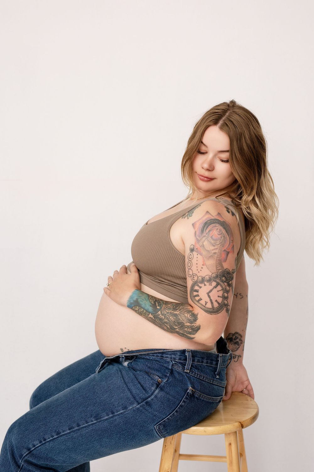 Pregnant person in brown tank top and jeans poses on wooden stool against white background, showing tattooed arms.