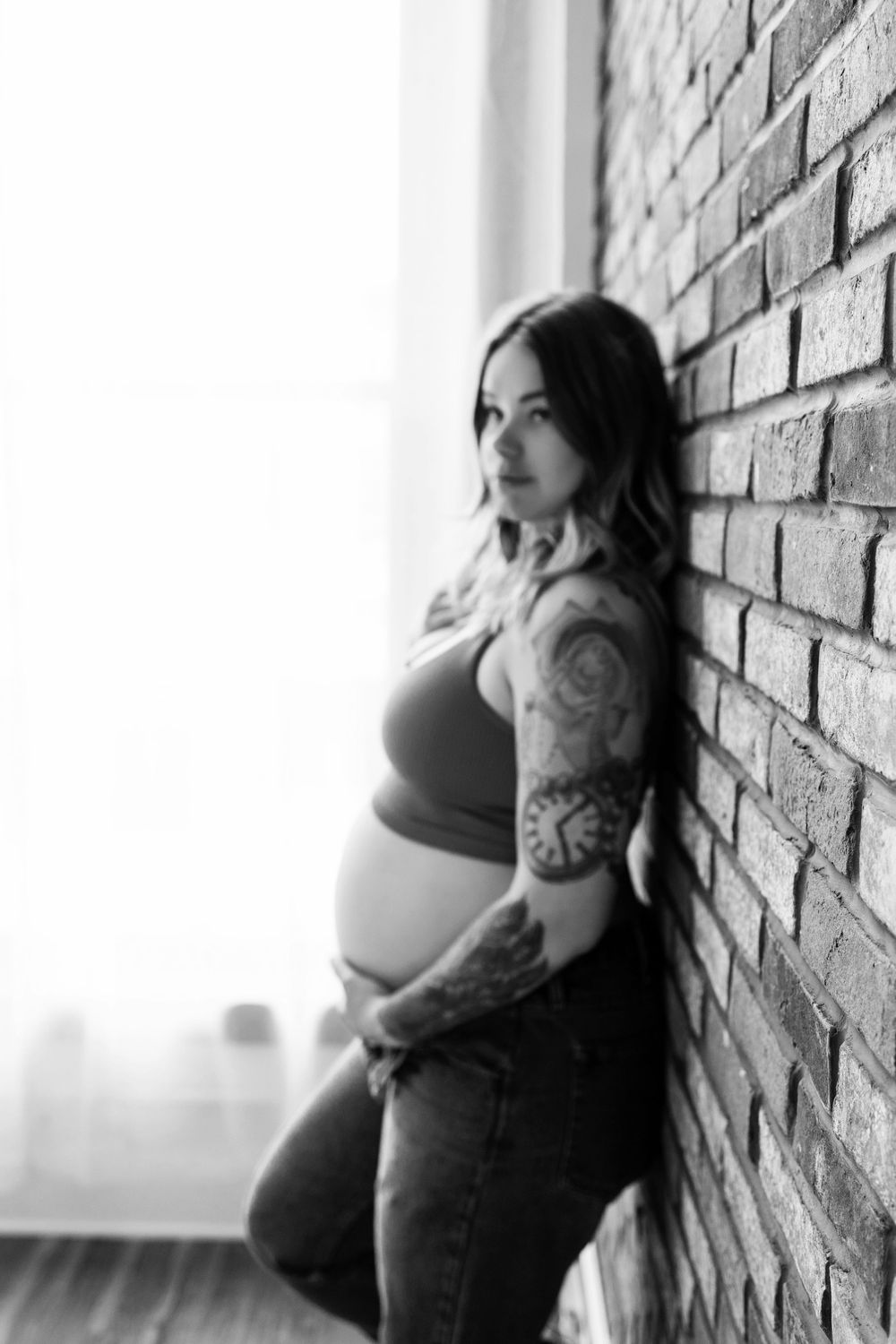 Black and white photo of an expectant mother in casual attire leaning against a brick wall. Watertown Wisconsin maternity photography.