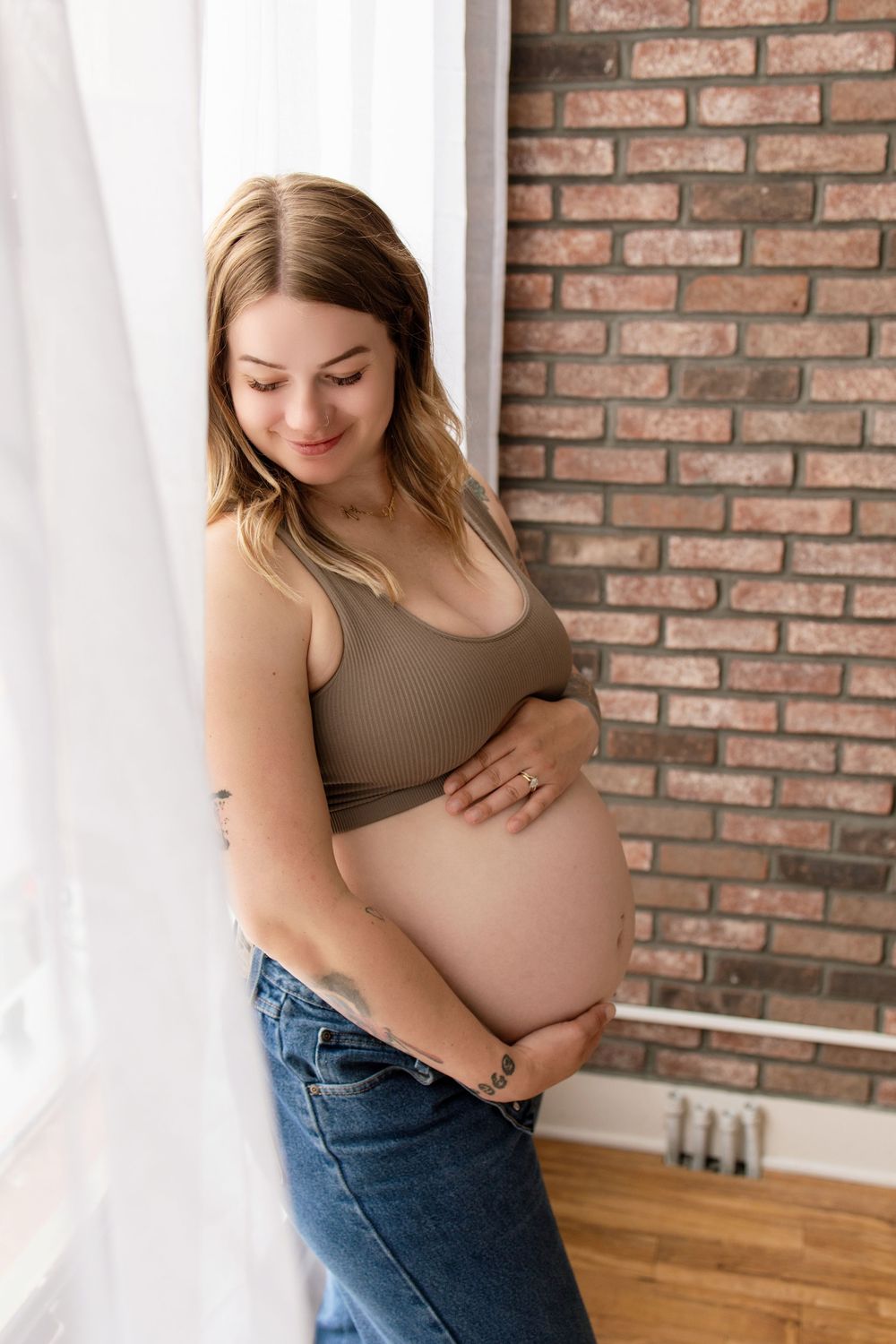 Pregnant woman in brown top and jeans cradles her belly while standing against exposed brick wall in natural light. Maternity Photography