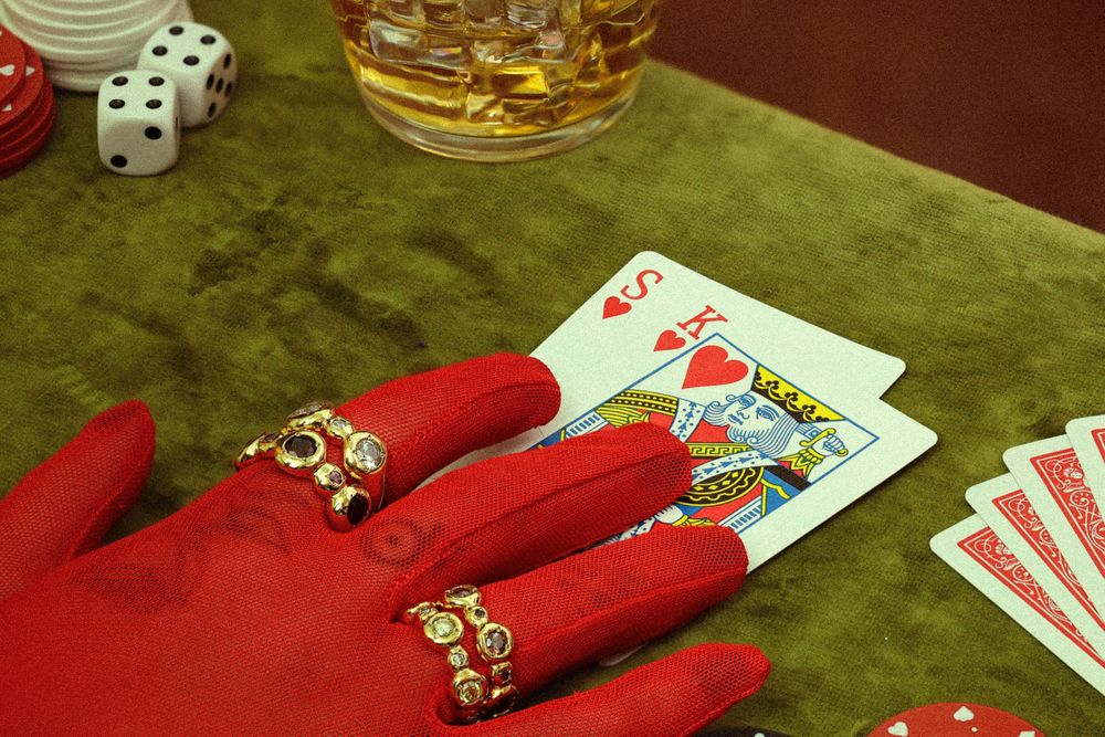 Playing cards and dice on green felt table with red glove wearing gold rings and glass of whiskey visible.