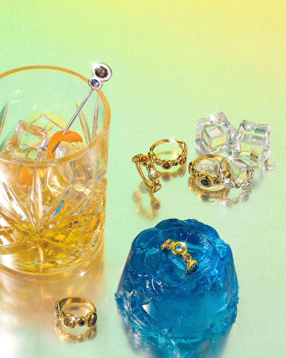 Decorative gold crown rings are displayed with ice cubes and cocktails against a gradient yellow-green background.