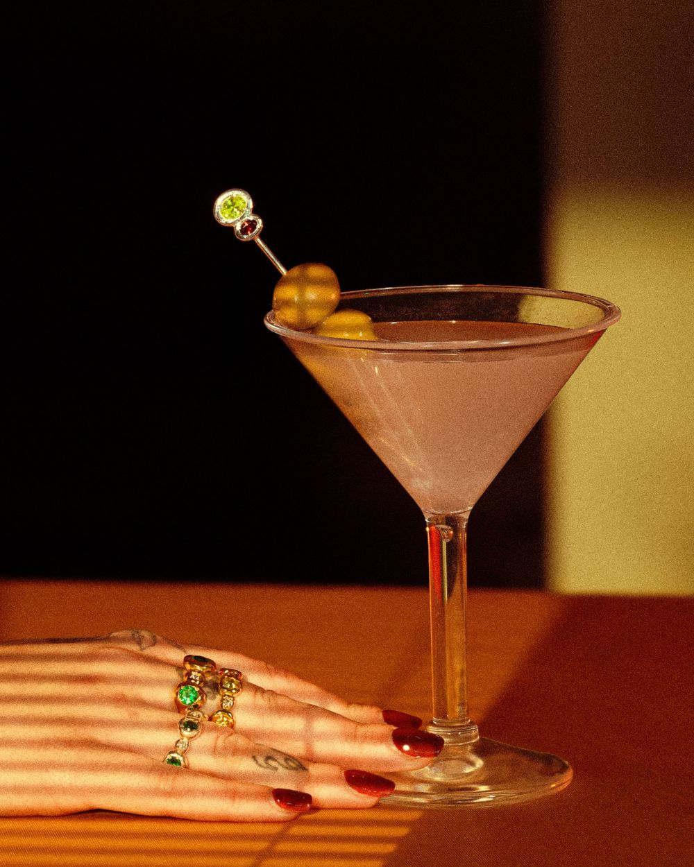 A pink martini cocktail with an olive garnish sits on a wooden surface with a hand wearing rings visible in the background.