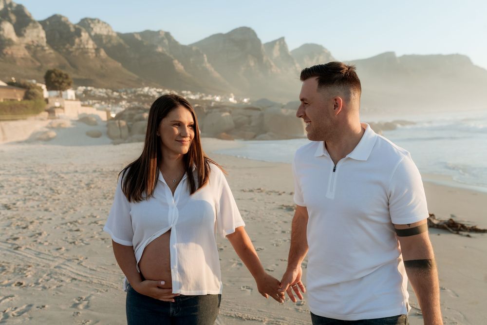 A couple in white shirts shares romantic moments on a scenic beach with mountains in the background at sunset.
