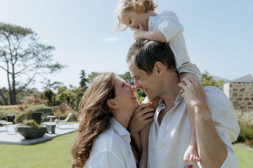 Happy family with young child outdoors, parents smiling at toddler sitting on father's shoulders in sunny garden.