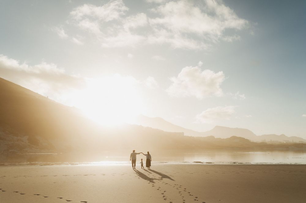 Two silhouettes walk along a misty beach at sunset with mountains and dramatic clouds in the background.