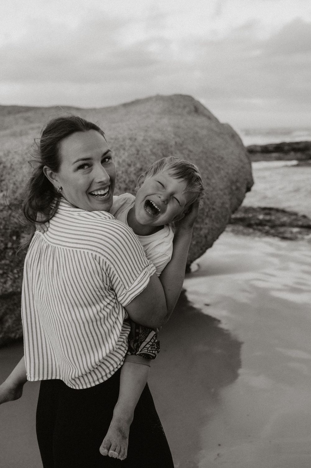 Black and white photo showing a joyful moment at the beach with a striped shirt and playful expression against rocky backdrop.