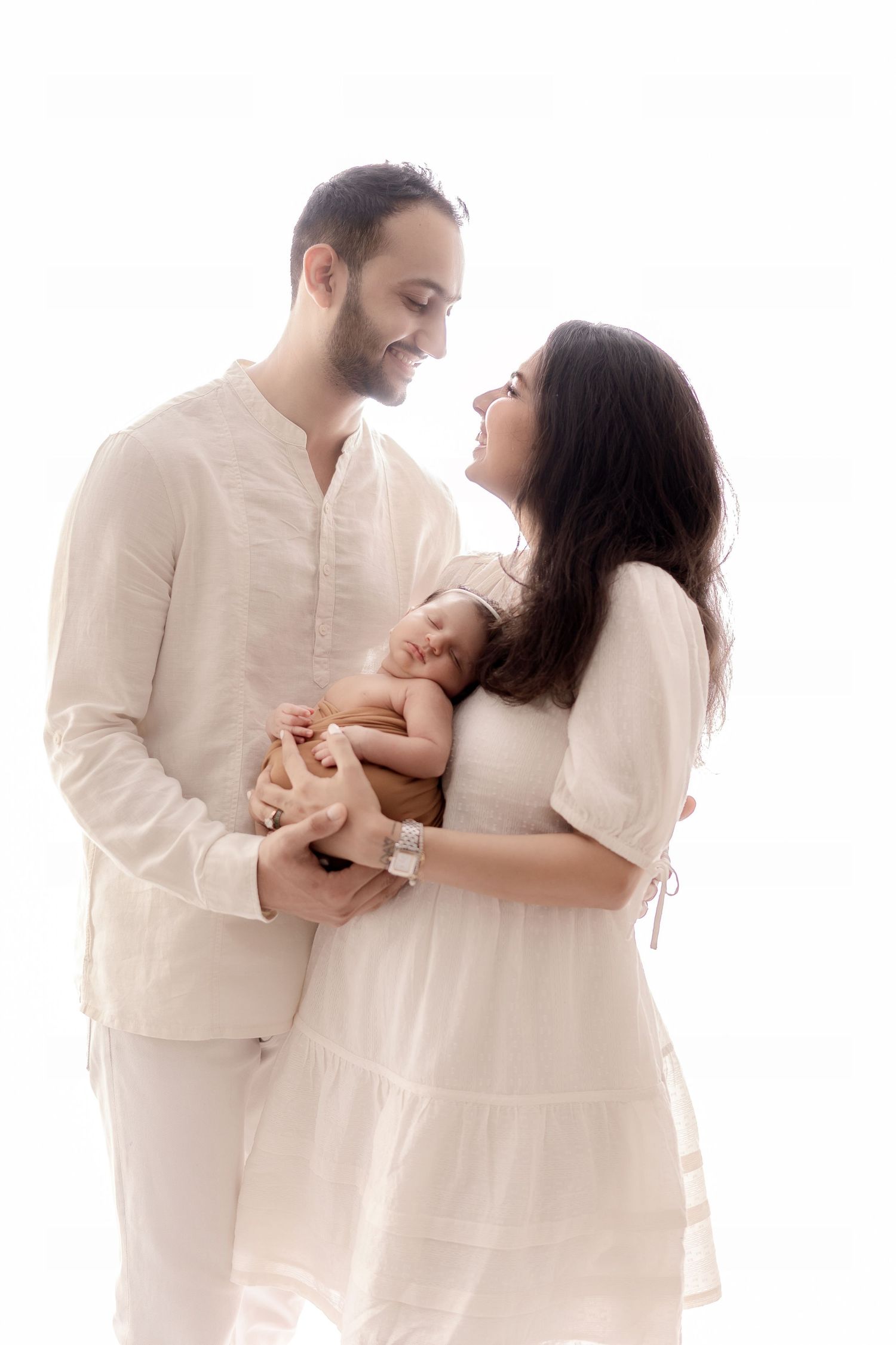 Family portrait in matching white outfits with newborn against bright background.