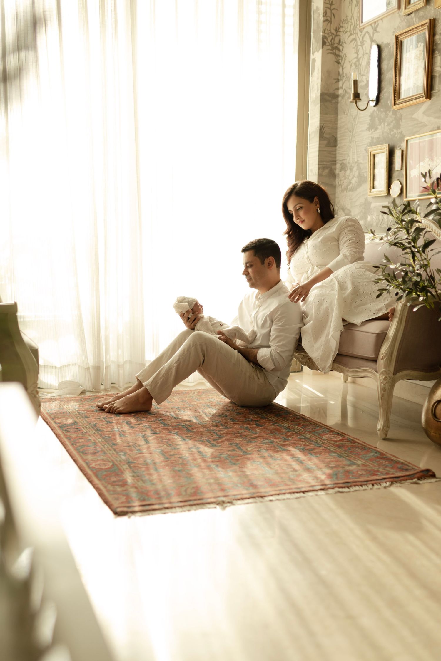 A couple in white outfits relaxing together on a patterned rug in a bright sunlit room with house plants and framed art.