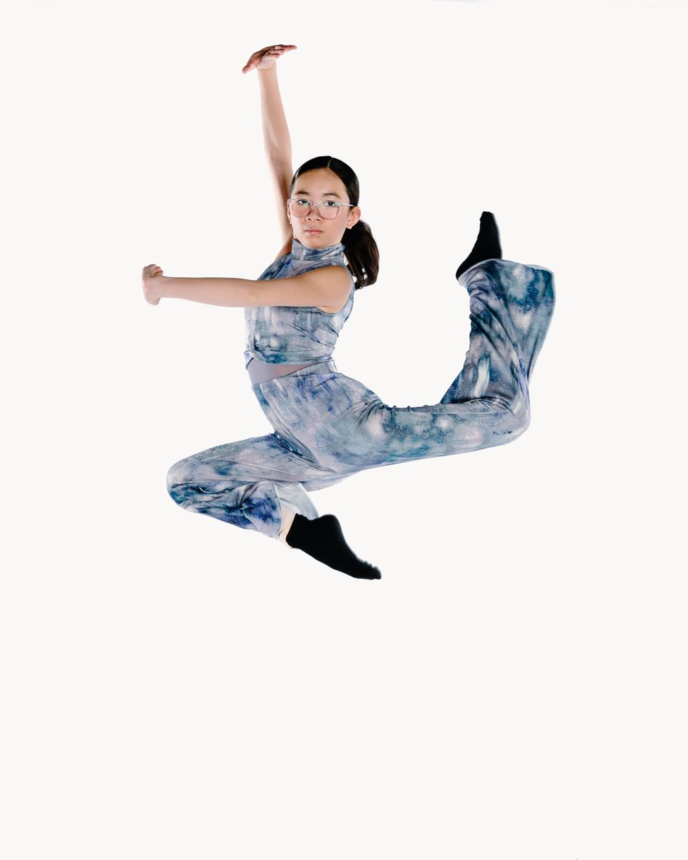 Dancer wearing blue tie-dye outfit performs dynamic leap against white background.