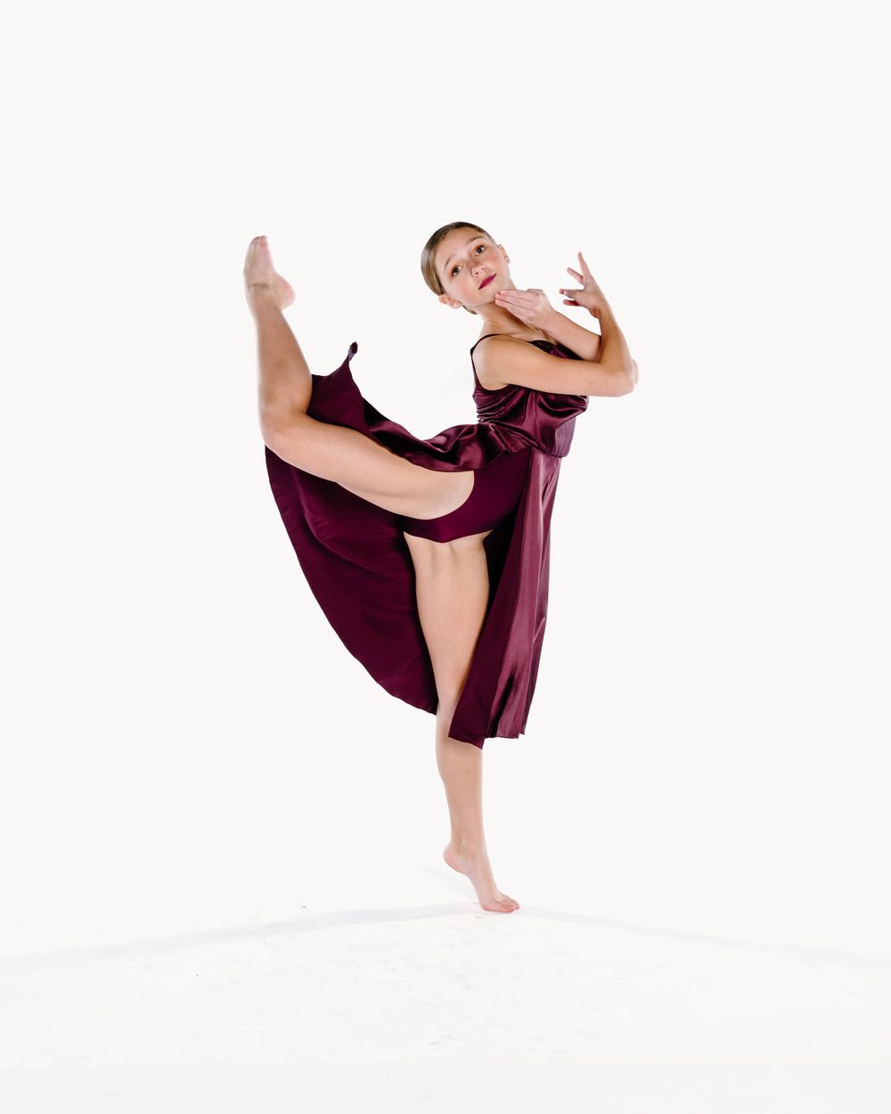 A ballet dancer in a flowing burgundy dress performs a graceful high kick movement against a white background.