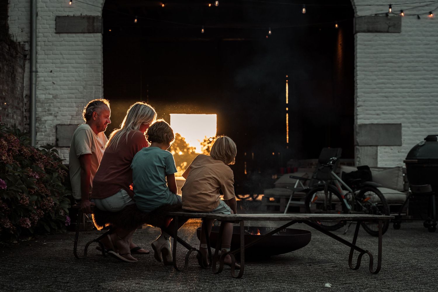 People gathered at a picnic table under string lights at dusk near a brick building entrance.