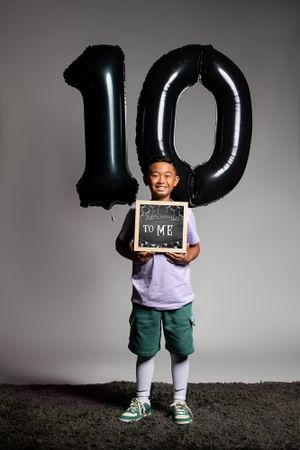 A young student stands with large black number 10 balloons and holds a small chalkboard sign against a grey background.