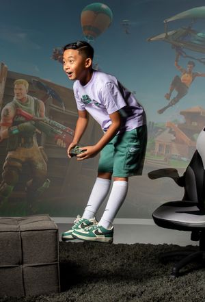 A young player in a white shirt and green shorts poses in motion against a surreal background with hot air balloons.