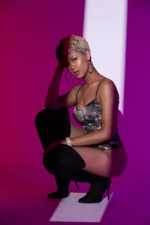 Fashion model wearing black boots and patterned bodysuit poses on pink background with dramatic lighting.