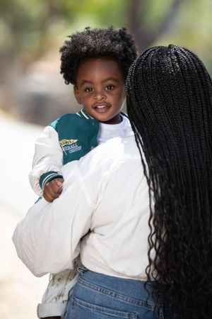 Child wearing a green varsity jacket and white shirt smiles while being held outdoors.