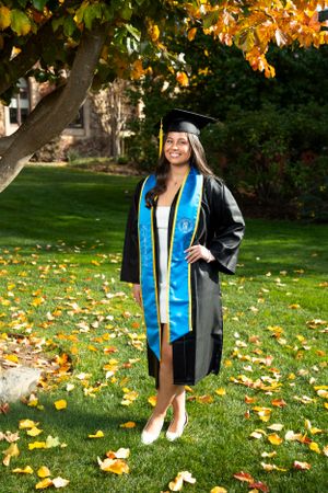 Graduate in cap and gown stands on autumn lawn with fallen leaves under tree with changing orange foliage.