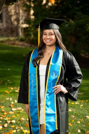 Graduate wearing traditional black cap and gown with bright blue stole poses outdoors on autumn day with fallen leaves.