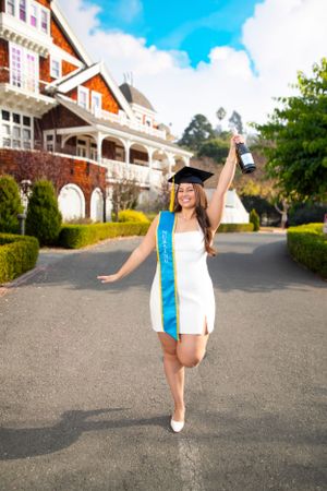 A graduate in white dress and blue sash celebrates joyfully outside a grand Victorian-style mansion on a sunny day.