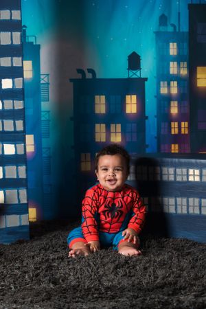 A toddler in a Spider-Man costume sits on a carpeted floor against a cityscape backdrop with blue lighting.