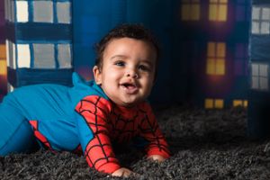 Toddler wearing Spider-Man costume crawls on gray carpet with colorful window lighting in background.