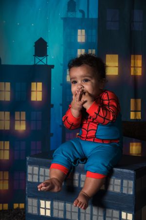 Toddler dressed as Spider-Man sits on ledge against cityscape backdrop in studio photo setting.
