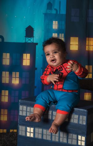 A toddler wearing a Spider-Man outfit sits on a prop with illuminated city buildings in the background.
