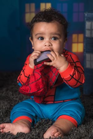 A baby wearing a Spider-Man costume sits on a dark carpet while chewing on a toy with colorful windows in the background.