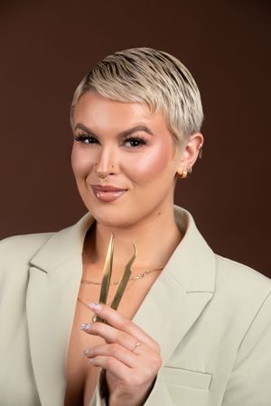 Professional portrait of someone with short platinum blonde hair wearing a cream blazer holding makeup brushes against brown backdrop.