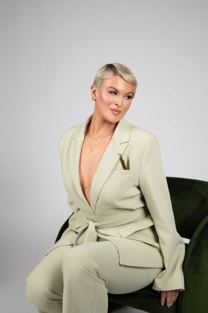 A person in a light beige pantsuit sits confidently on a dark green velvet chair against a gray backdrop.