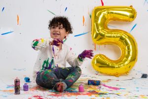 Creative portrait of child sitting with paint splashes and a gold number 5 balloon against white background for birthday shoot.
