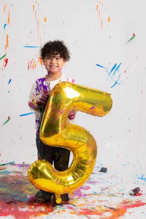 A large gold number 5 balloon held in a colorful paint splattered setting with vibrant confetti and blue strokes. Birthday photos