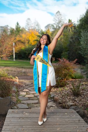 Graduate wearing blue and white graduation stole strikes joyful pose on wooden pathway during autumn photoshoot.
