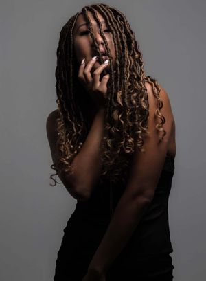 Artistic portrait against gray backdrop featuring figure with long dreadlocks in dramatic lighting and black top.