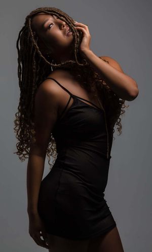Artistic portrait of model in black dress with long braids striking dramatic pose against gray background.