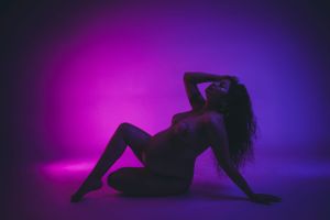 Silhouette of a feminine figure in a sensual pose against a vibrant purple and blue backlit background. maternity photoshoot
