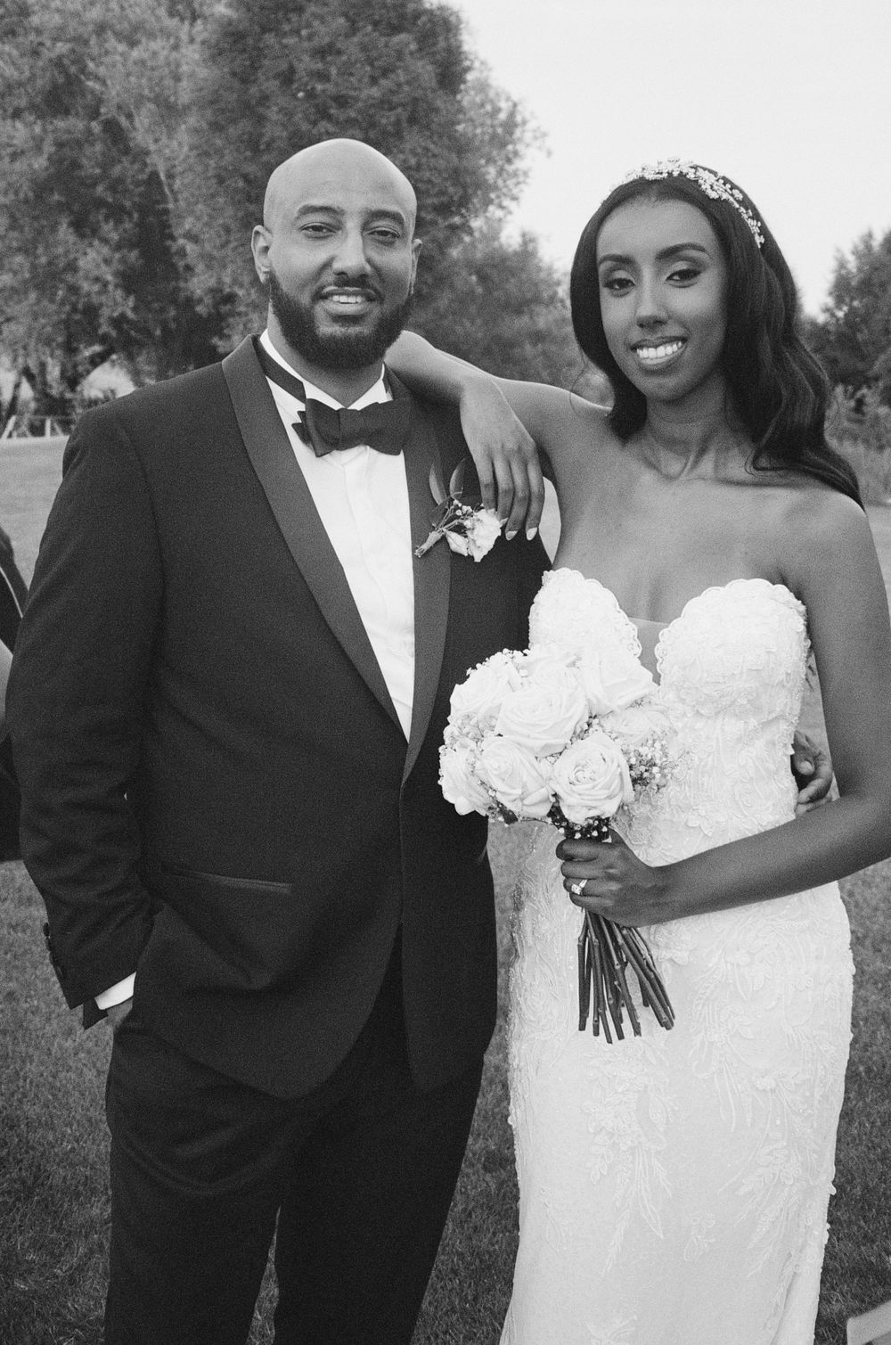 Black and white wedding photo of a smiling bride in strapless gown holding bouquet beside groom in tuxedo outdoors.