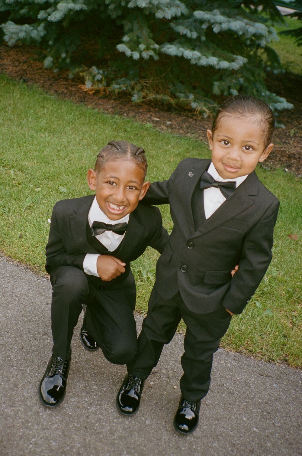 Two young boys in matching black tuxedos and bow ties pose together outdoors on a pathway.