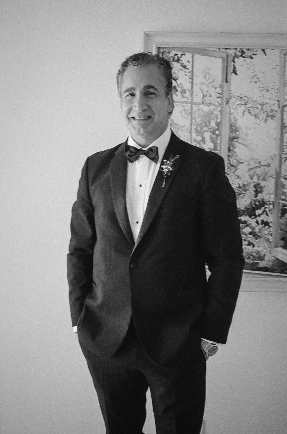 A man in a black tuxedo with bow tie and boutonniere smiles in a black and white wedding photo.
