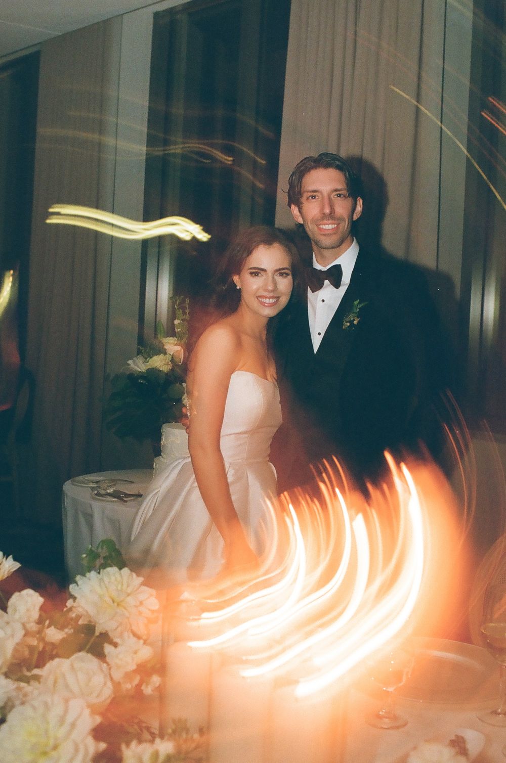 Bride and groom smiling at wedding reception with bokeh light streaks and floral arrangements in foreground.