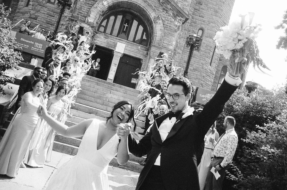 Joyful newlyweds celebrate outside a historic stone church, groom raising arms triumphantly, bride holding bouquet, wedding party nearby.